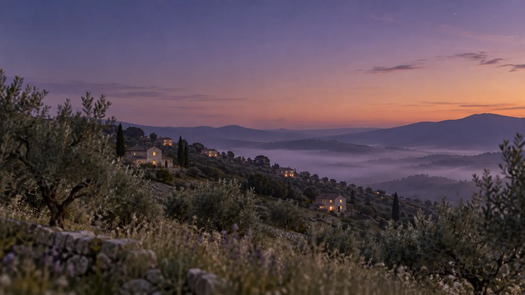 A hillside at dusk with a few lit windows scattered across the slope above a misty valley